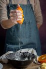 Couper la main d'une femme méconnaissable dans un tablier serrant de l'orange fraîche coupée juteuse sur un bol tout en préparant la pâte à table — Photo de stock