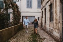 Alegre pareja joven en ropa casual elegante cogidas de la mano y sonriendo mientras camina por la antigua calle estrecha de la ciudad - foto de stock
