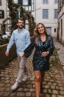 Jovem casal alegre em roupas casuais elegantes de mãos dadas e sorrindo enquanto caminhava na rua estreita velha na cidade — Fotografia de Stock