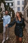 Cheerful young couple in stylish casual clothes holding hands and smiling while walking on old narrow street in city — Stock Photo