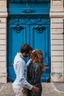 Вид сбоку счастливой молодой пары в повседневной одежде, обнимающей и целующейся, стоя перед старым каменным зданием с голубыми дверями на городской улице — стоковое фото