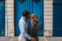 Вид сбоку счастливой молодой пары в повседневной одежде, обнимающей и целующейся, стоя перед старым каменным зданием с голубыми дверями на городской улице — стоковое фото