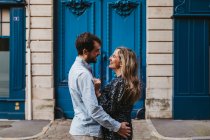 Side view of happy young couple in casual clothes hugging looking at each other while standing against aged stone building with blue doors on city street — Stock Photo