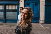 Glückliche junge Frau blickt in stilvollem Outfit in die Kamera, gefolgt von lächelndem Freund, der während eines romantischen Urlaubs in Frankreich auf der Stadtstraße mit altem Gebäude im Hintergrund spaziert — Stockfoto