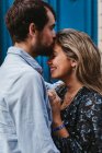 Вид збоку щасливої молодої пари в повсякденному одязі обіймає і цілує, стоячи проти старого кам'яного будинку з блакитними дверима на вулиці міста — стокове фото