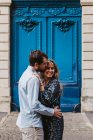 Vista lateral de la feliz pareja joven en ropa casual abrazos y besos, mientras que de pie contra el edificio de piedra envejecida con puertas azules en la calle de la ciudad - foto de stock