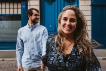Happy young woman looking at camera in stylish outfit followed by smiling boyfriend walking on city street with old building in background during romantic holidays in France — Stock Photo