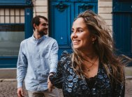 Happy young woman in stylish outfit followed by smiling boyfriend walking on city street with old building in background during romantic holidays in France — Stock Photo