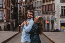 Vista lateral de la alegre pareja joven en ropa casual abrazándose y mirándose con amor mientras están de pie en la calle contra el viejo edificio durante las vacaciones románticas en Francia - foto de stock