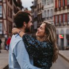 Вид сбоку на веселую молодую пару в повседневной одежде, обнимающую и смотрящую друг на друга с любовью, стоя на улице напротив старого здания во время романтического отдыха во Франции — стоковое фото