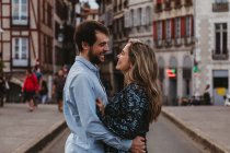 Side view of cheerful young couple in casual wear embracing and looking at each other with love while standing on street against old building during romantic holidays in France — Stock Photo