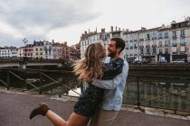 Vue latérale d'un jeune couple heureux et affectueux embrassant et riant debout sur un remblai de pierre près d'une rivière avec de vieux bâtiments en arrière-plan dans la ville de Bayonne en France — Photo de stock