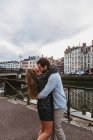 Vista lateral de jovens felizes casal afetuoso abraçando e beijando enquanto estava de pé em aterro de pedra perto do rio com edifícios antigos no fundo na cidade de Bayonne, na França — Fotografia de Stock