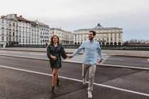 Щаслива молода романтична пара в стильному одязі сміється і тримається за руки, перетинаючи міст з історичними будівлями на задньому плані під час поїздки в місто Байонна (Франція). — стокове фото