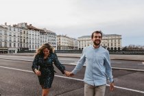 Happy young romantic couple in stylish clothes laughing and holding hands while crossing bridge with historic buildings in background during city tour in Bayonne in France — Stock Photo