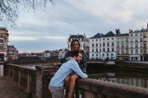 Jovem feliz sentada na cerca de pedra velha e abraçando namorado afetuoso enquanto passar a noite de verão juntos em Bayonne — Fotografia de Stock