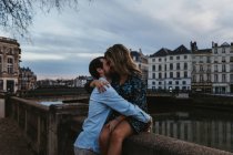 Jovem feliz sentada na cerca de pedra velha abraçando e beijando namorado afetuoso enquanto passam a noite de verão juntos em Bayonne — Fotografia de Stock