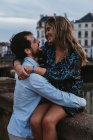 Feliz joven hembra sentada en una vieja valla de piedra y abrazando a un novio cariñoso mientras pasan la noche de verano juntos en Bayona - foto de stock