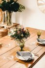 Strauß aus verschiedenen Blumen und grünen Pflanzenzweigen in einer Vase mit Wasser auf einem Holztisch für eine Mahlzeit — Stockfoto