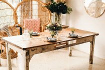 De arriba ramo de flores diversas y ramitas de plantas verdes en jarrón con agua en una mesa de madera para una comida con hermosa silla de ratán diseñada en el fondo - foto de stock