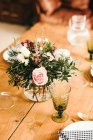 From above bouquet of miscellaneous flowers and green plant twigs in vase with water on a wooden table set for a meal — Stock Photo