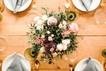 De dessus vue bouquet de fleurs diverses et brindilles de plantes vertes dans un vase avec de l'eau sur une table en bois ensemble pour un repas — Photo de stock