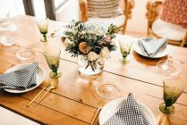 De acima mencionado buquê de flores diversas e ramos de fábrica verdes no vaso com a água em uma mesa de madeira posta para uma refeição — Fotografia de Stock