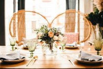Bouquet of miscellaneous flowers and green plant twigs in vase with water on a wooden table set for a meal with beautiful designed rattan chair on the background — Stock Photo