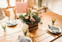 From above bouquet of miscellaneous flowers and green plant twigs in a wooden box on a timber table set for a meal with beautiful designed rattan chair on the background — Stock Photo