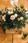 Сверху букет разноцветных цветов и зеленых цветочных веточек в деревянной коробке на деревянном столе, накрытом на обед — стоковое фото
