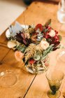 Dall'alto mazzo di fiori vari e ramoscelli di pianta verdi in vaso con acqua su una tavola di legno apparecchiata per un pasto — Foto stock