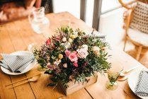 Von oben Bouquet von verschiedenen Blumen und grünen Pflanzenzweigen in einer Holzkiste auf einem Holztisch gedeckt für eine Mahlzeit mit schön gestalteten Rattanstuhl auf dem Hintergrund — Stockfoto