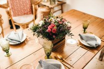 De arriba ramo de flores diversas y ramitas de plantas verdes en una caja de madera en una mesa de madera para una comida con hermosa silla de ratán diseñada en el fondo - foto de stock