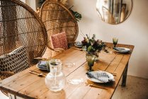 From above bouquet of miscellaneous flowers and green plant twigs in vase with water on a wooden table set for a meal with beautiful designed rattan chair on the background — Stock Photo