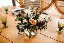 De cima buquê de flores diversas e ramos de plantas verdes em vaso com água em uma mesa de madeira para uma refeição com bela cadeira de vime projetada no fundo — Fotografia de Stock