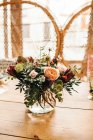 Ramillete de flores diversas y ramitas de plantas verdes en jarrón con agua en una mesa de madera para una comida con hermosa silla de ratán diseñada en el fondo - foto de stock