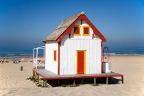 Pequeña casa de campo blanca con puerta roja y ventanas situadas en la playa contra el cielo azul sin nubes en el día soleado - foto de stock