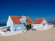 Piccola casa di campagna a strisce bianche e blu con recinzione bianca situata al mare contro il cielo blu senza nuvole nella giornata di sole — Foto stock