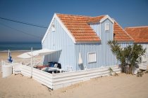 Petite maison de campagne à bandes blanches et bleues avec clôture blanche située au bord de la mer contre un ciel bleu sans nuages par temps ensoleillé — Photo de stock