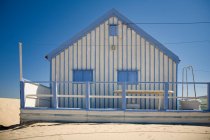 Piccola casa di campagna a strisce bianche e blu con recinzione bianca situata al mare contro il cielo blu senza nuvole nella giornata di sole — Foto stock