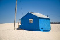 Маленький голубой дом с потрепанными стенами расположен на песчаном побережье с голубым небом на заднем плане в солнечный день — стоковое фото