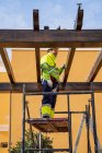 Vue de dessous du technicien masculin en tenue de travail debout sur un échafaudage et se préparant à installer un panneau solaire sur une construction en bois — Photo de stock