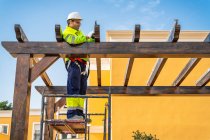 Dal basso vista laterale di tecnico maschile in abbigliamento da lavoro in piedi su ponteggi e preparazione per l'installazione di pannello solare su costruzione in legno — Foto stock