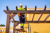 Dal basso vista laterale di tecnico maschile in abbigliamento da lavoro in piedi su ponteggi e preparazione per l'installazione di pannello solare su costruzione in legno — Foto stock