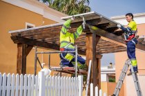 Grupo de trabalhadores em uniforme e capacetes instalando painéis fotovoltaicos no telhado de construção de madeira perto da casa — Fotografia de Stock