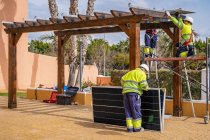 Група робітників у формі та шоломах встановлюють фотоелектричні панелі на даху дерев'яного будівництва біля будинку — стокове фото