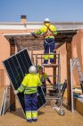 Grupo de trabajadores en uniforme y cascos instalando paneles fotovoltaicos en techo de construcción de madera cerca de casa - foto de stock