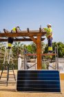 Grupo de trabajadores en uniforme y cascos instalando paneles fotovoltaicos en techo de construcción de madera cerca de casa - foto de stock