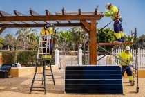Group of workers in uniform and helmets installing photovoltaic panels on roof of wooden construction near house — Stock Photo