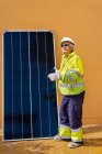 Побочный обзор опытного мужчины-техника в форме и шлеме, стоящего рядом с солнечной панелью возле желтого здания во время работы по установке системы возобновляемой энергии — стоковое фото